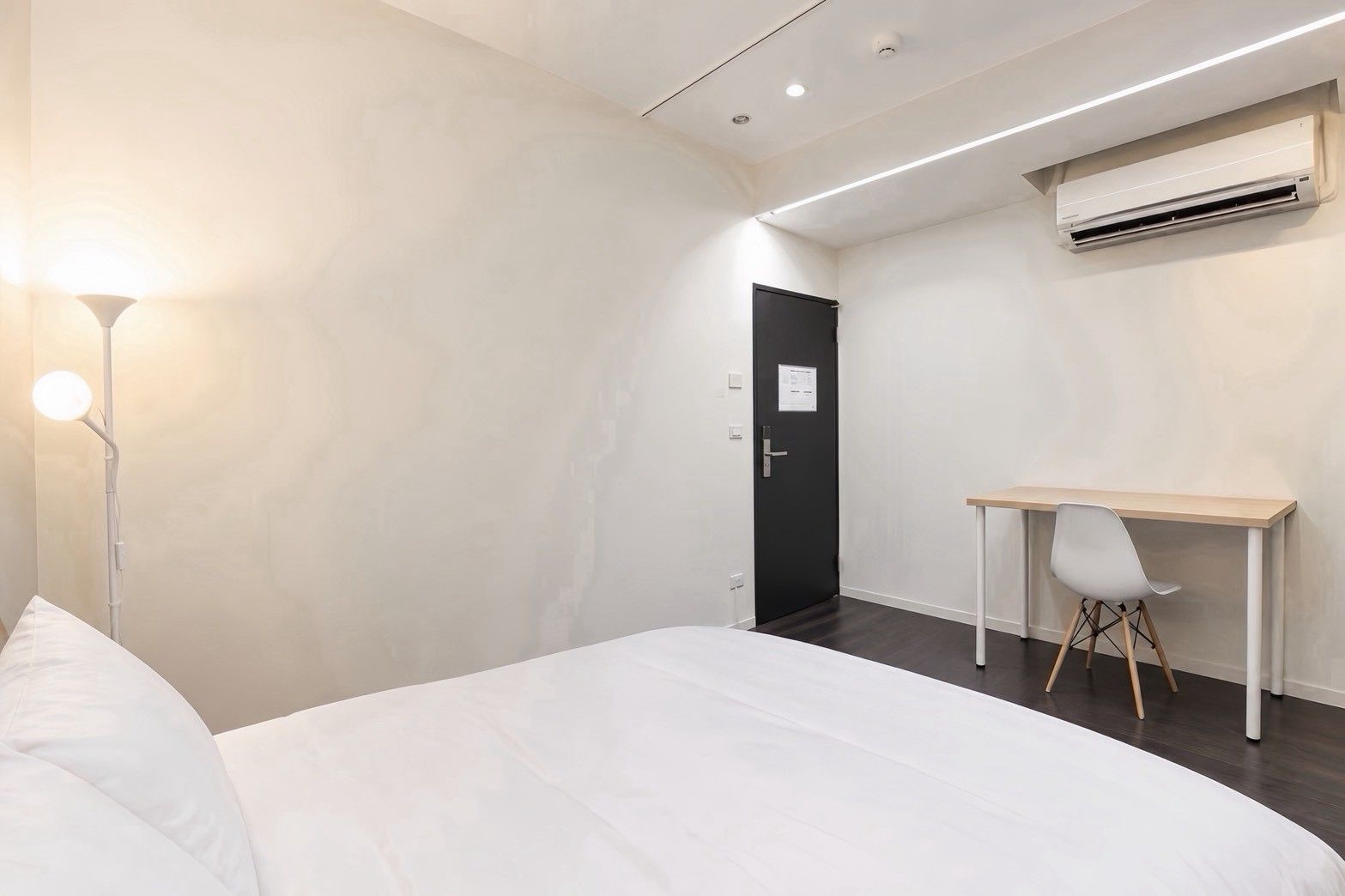 A hotel room with a bed, a desk, a chair, an air conditioning unit, and a door.