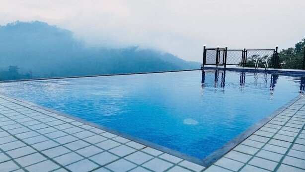 Une magnifique piscine à débordement d’un bleu profond, avec une terrasse carrelée, entourée d’une clôture et offrant une vue imprenable sur des montagnes brumeuses.