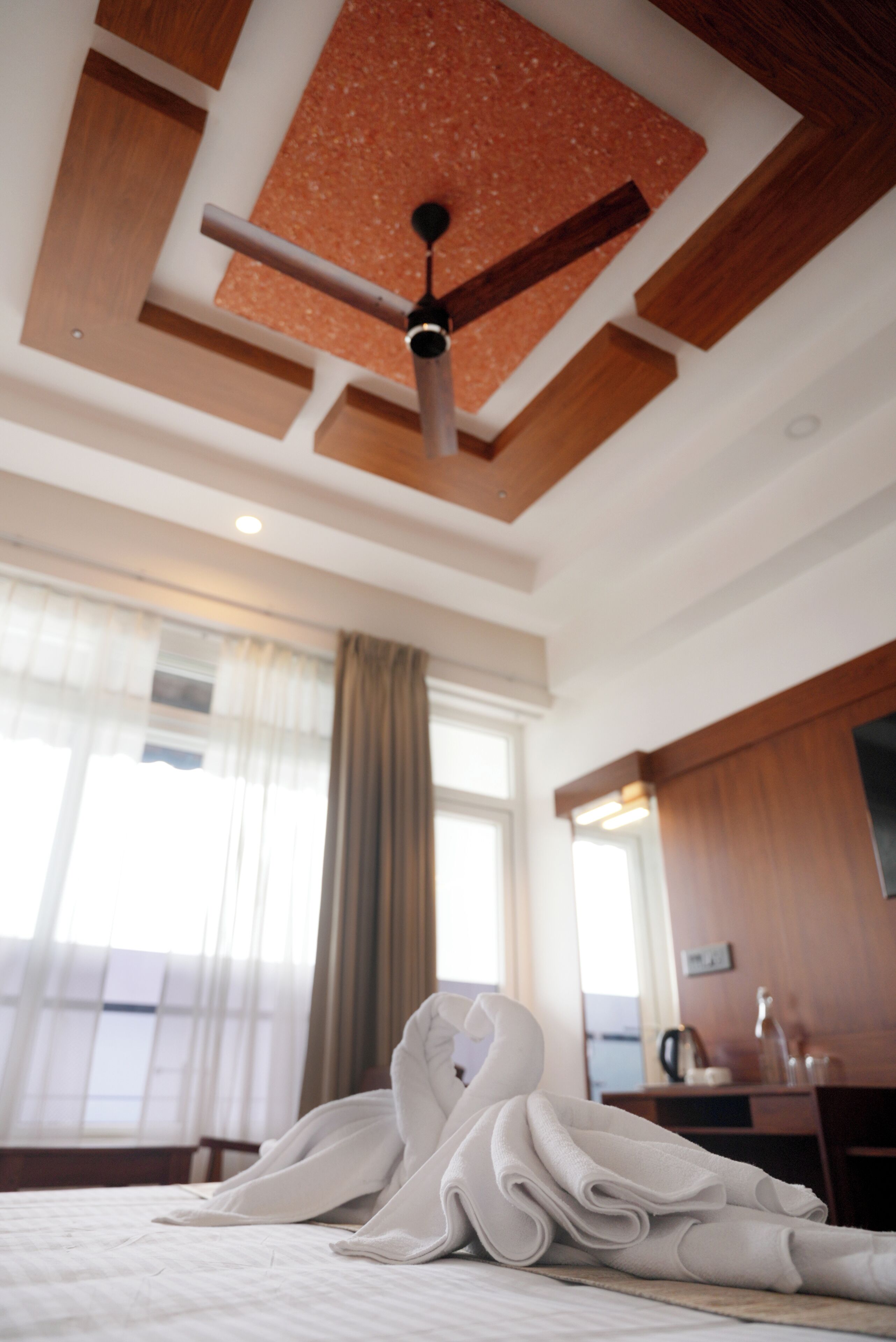A hotel room with a ceiling fan, a bed with neatly folded white towels, a wooden desk, and a window with curtains.