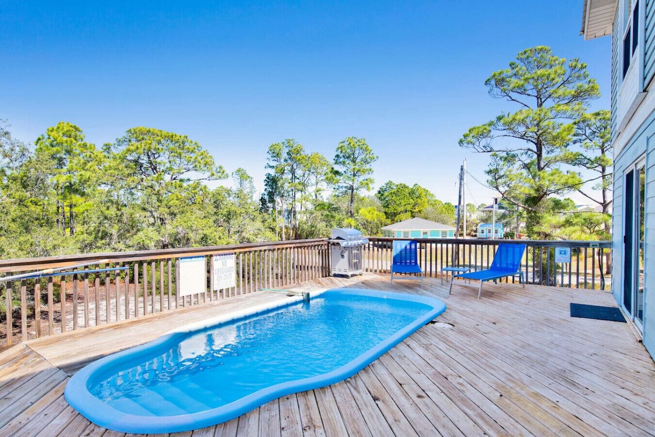 Piscina exterior en una terraza de madera, con un letrero que indica la presencia de un socorrista, rodeada de árboles y una casa.