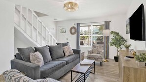 A modern living room with a grey sofa, a coffee table, and a TV. There is a staircase with white railings, a chandelier, and a window with blinds. - Sleeps 16 · Private Pool · 3 Blocks to Beach (Gulf Shores)