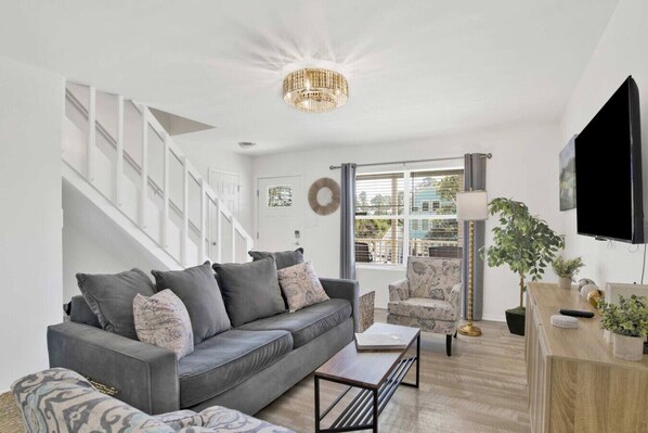 A modern living room with a grey sofa, a coffee table, and a TV. There is a staircase with white railings, a chandelier, and a window with blinds.