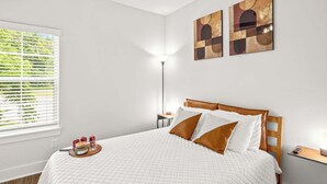 Ein ordentlich bezogenes Bett mit weißer gesteppter Bettdecke und zwei braunen Kissen, ein Nachttisch mit Lampe und ein Fenster mit Jalousien.