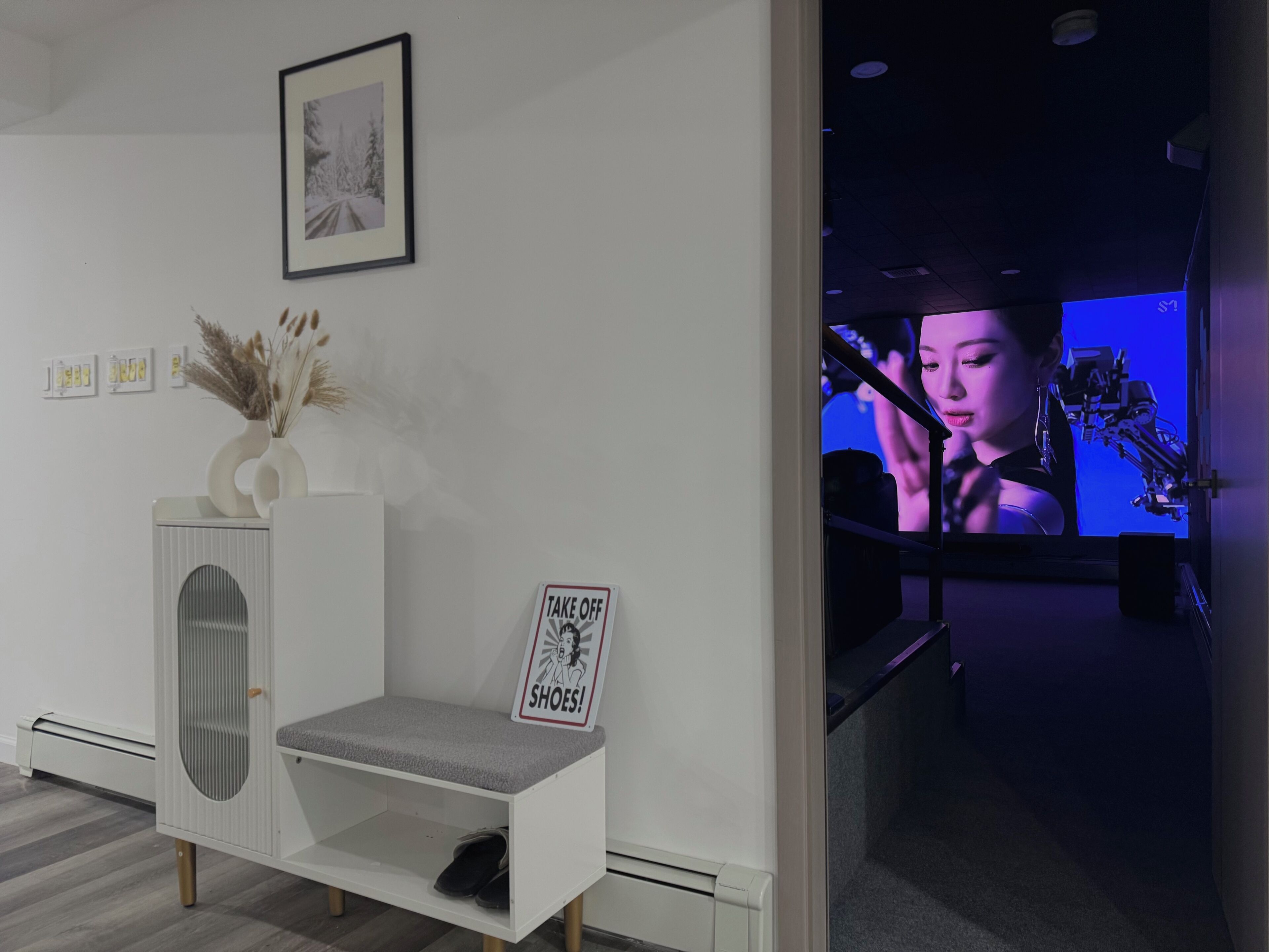 Una habitación con un armario blanco, un cuadro enmarcado, un letrero que dice '¡Quítese los zapatos!' y una pantalla de televisión que muestra a una persona.