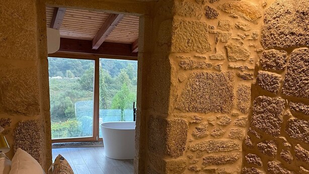 Ein Schlafzimmer mit Steinwand, einem Bett mit weißer Bettwäsche und Blickrichtung auf ein Badezimmer sowie auf Grünflächen draußen.