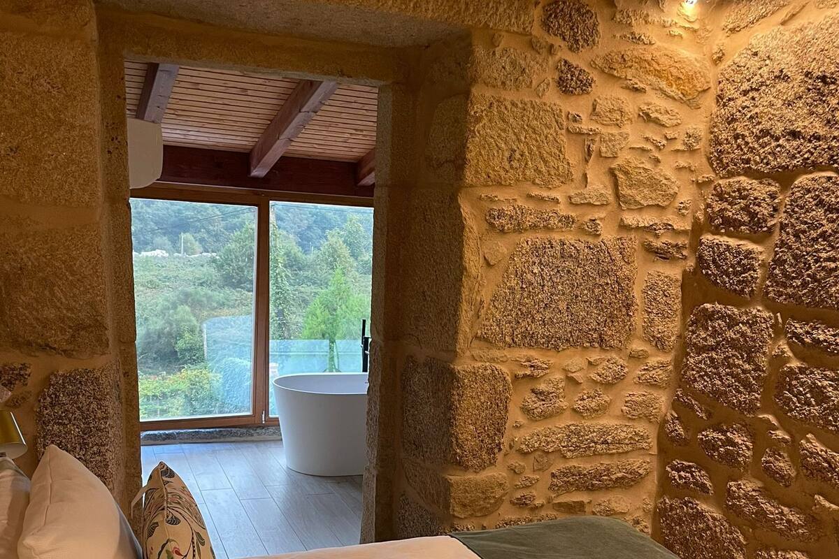 Une chambre avec un mur en pierre, un lit avec une literie blanche et une vue sur une salle de bain et la verdure à l’extérieur.