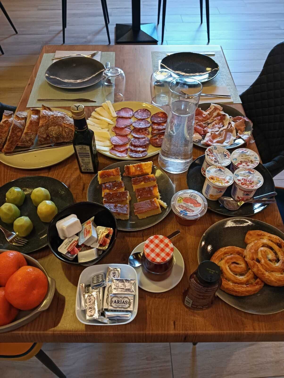 Una mesa servida con diversos alimentos, como pan, queso, jamón y pasteles.