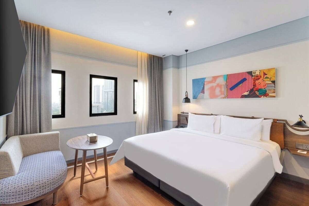 A hotel room with a large bed, a chair, a small table, and a painting on the wall.