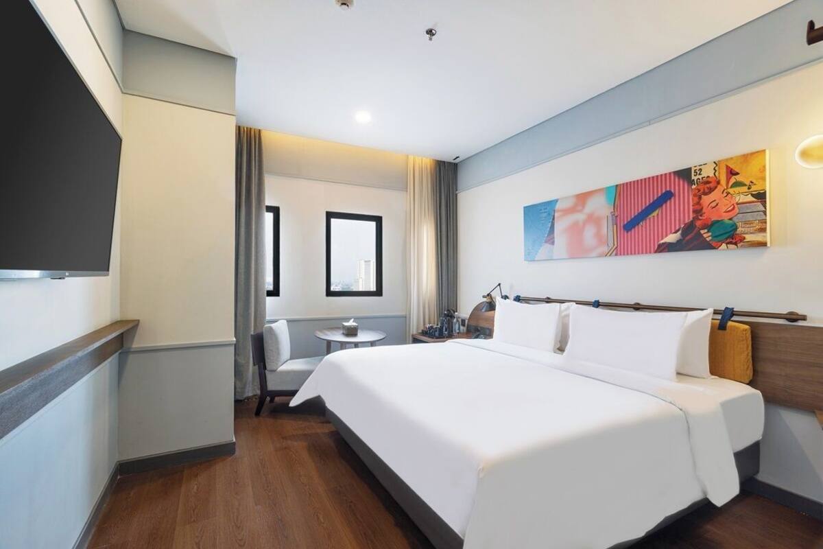 A hotel room with a large bed, a desk, and a chair. There is a window with curtains, a painting on the wall, and a television mounted on the wall.