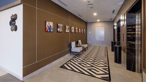Een moderne lobby met een vloerkleed met geometrisch patroon, een zithoek met twee stoelen en een rij kleurrijke abstracte schilderijen aan de muur.
