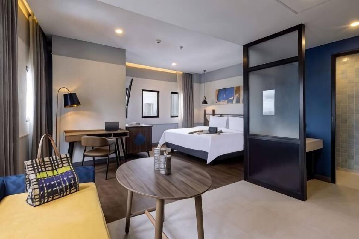 A hotel room with a bed, desk, chair, table, and a view of the city.