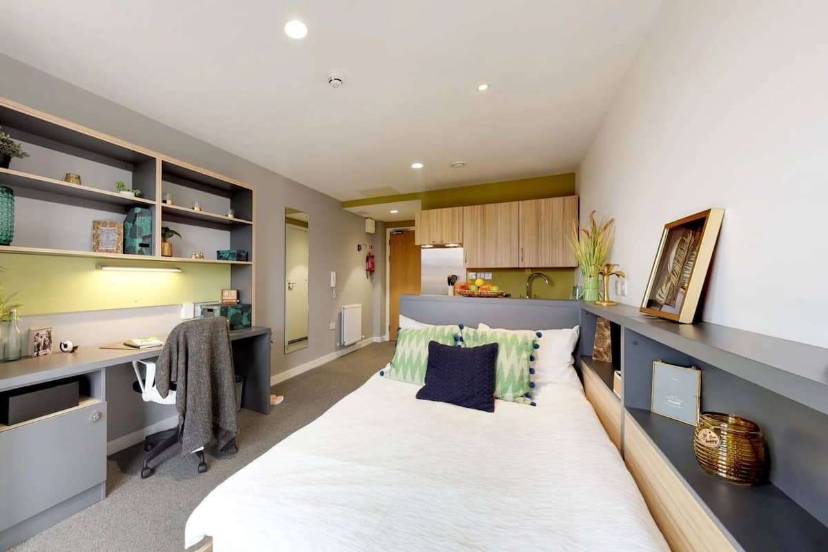 A modern hotel room with a bed, desk, and kitchenette.