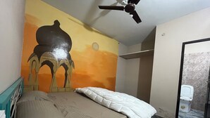 A bedroom with a bed, a ceiling fan, a mural on the wall, and a bathroom with a toilet visible through an open door. - Jungle by sturmfrei Jaipur (Jaipur)