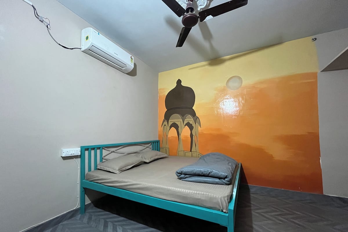 Un lit simple avec un cadre de couleur turquoise et une literie grise, dans une pièce dont le sol est recouvert d’un parquet à motifs en chevrons.