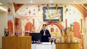 A hotel reception desk with a digital screen displaying the hotel name, a framed picture above the desk, and a decorative wall with patterns. - Deotel Inn Pithampur by JMC (Dr. Ambedkar Nagar)