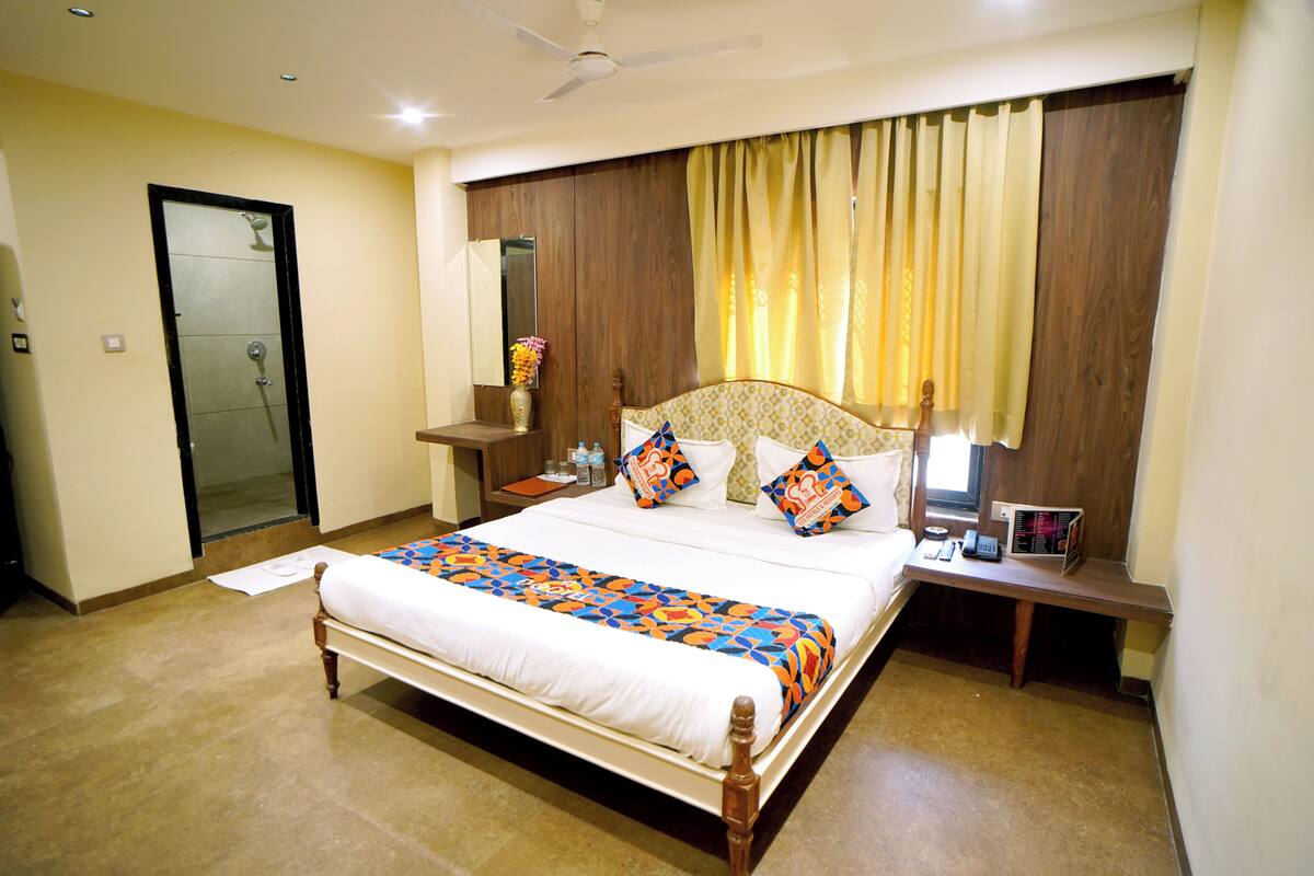 A hotel room with a large bed, a bedside table with a lamp, a nightstand, a mirror, a window with curtains, and a door.