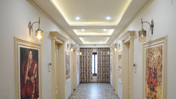 A hallway with patterned floor tiles, wall-mounted artwork, recessed ceiling lighting, and a view into a room with patterned curtains. - Deotel Inn Pithampur by JMC (Dr. Ambedkar Nagar)