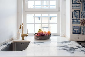 A kitchen with a marble countertop, a stainless steel sink, a fruit bowl, and a window with a view of buildings.