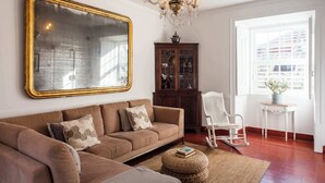 A living room with a large mirror, a chandelier, a wooden cabinet, a rocking chair, and a window with a view of buildings. - Central Ericeira Apartment with Lift (Ericeira)