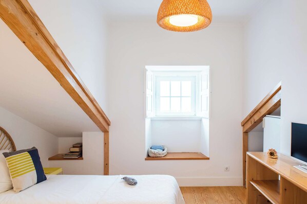 A bedroom with a bed, a window, a shelf with books, a TV, and a hanging light fixture.