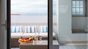 A balcony with a wicker table holding a fruit bowl, wine glasses, and a bottle. A white sofa with blue and white striped cushions is visible in the background. - Ocean View Penthouse with Rooftop Terrace! (Ericeira)