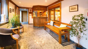 Een hotel lobby met een houten bankje, een kleine tafel en stoelen. Er staat een potplant en een lijst met een foto aan de muur.