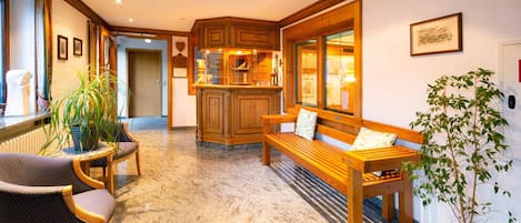 A hotel lobby with a wooden bench, a small table, and chairs. There is a potted plant and a framed picture on the wall.