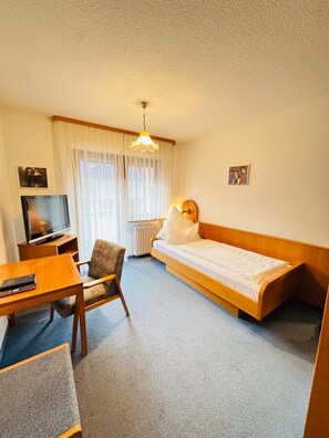 A compact hotel room with a single bed, a small table with a chair, a television, and a window with sheer curtains. - Hotel Grünwinkel (Ortenberg)