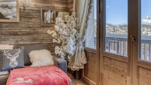 A cozy room with wooden walls, a large window with a view of a snowy landscape, a plush sofa with decorative pillows, and a vase of white flowers. - Central Megève Apartment - 4 Guests - Pets Allowed (Megève)