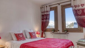 A bedroom with a bed, red and white bedding, a nightstand, a lamp, a window with curtains, and a view of mountains. - Central Megève Apartment - 4 Guests - Pets Allowed (Megève)