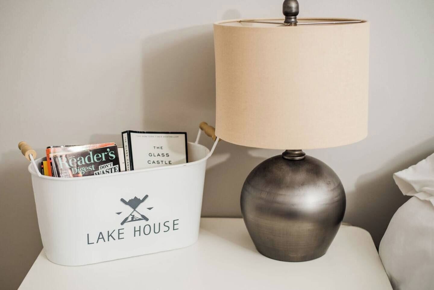 Una mesita de noche con una lámpara y una cesta blanca con la etiqueta “LAKE HOUSE” que contiene libros y revistas.