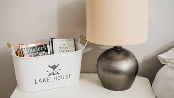 Una mesita de noche con una lámpara y una cesta blanca con la etiqueta “LAKE HOUSE” que contiene libros y revistas.