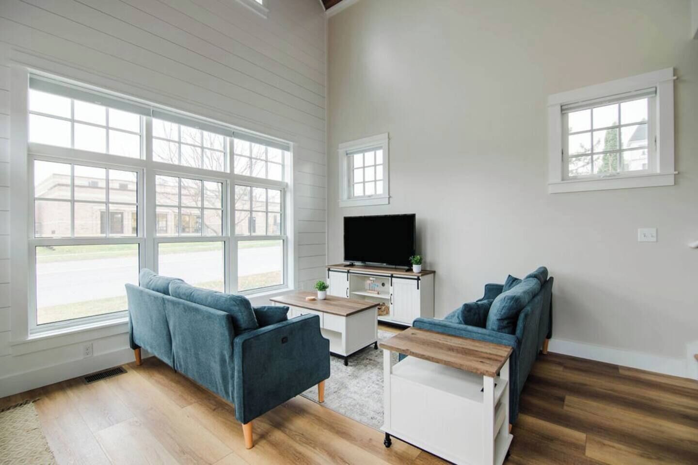 A spacious living room with large windows, a flat-screen TV, two blue sofas, and a wooden coffee table.