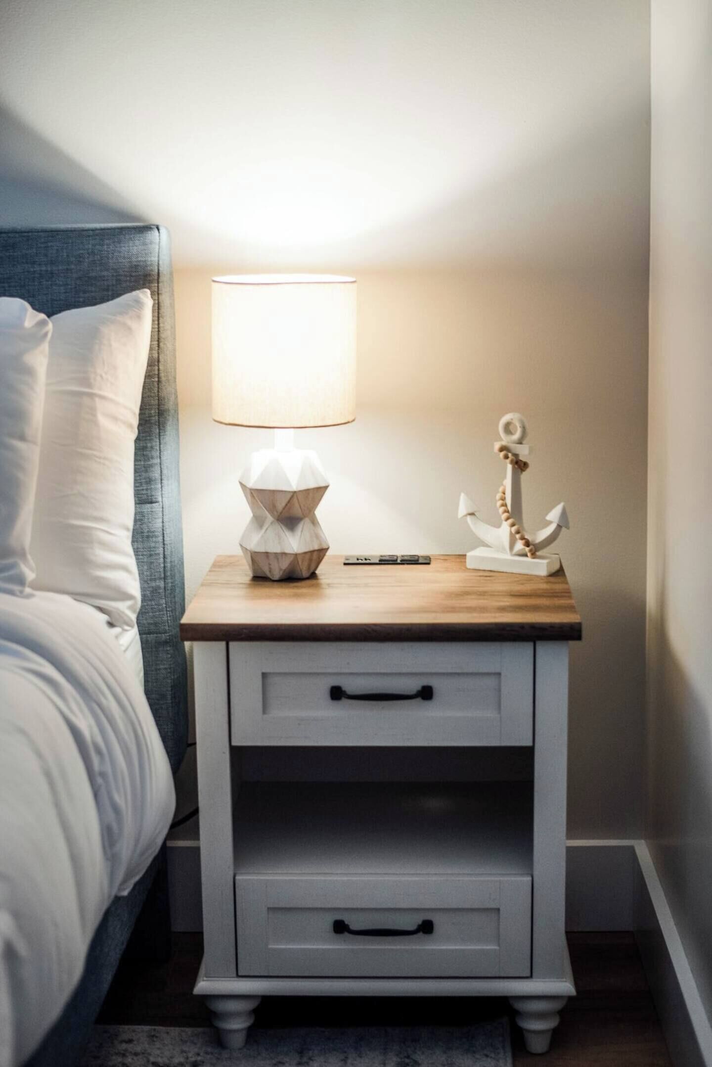 A neatly made bed with a headboard, a bedside lamp, a small wooden nightstand with drawers, and a decorative anchor.