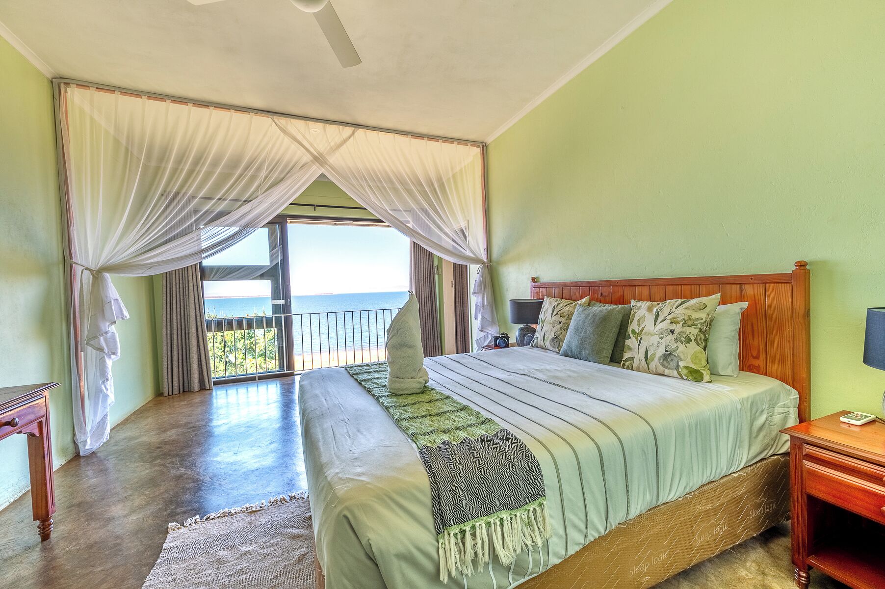 Een slaapkamer met een groot bed, een houten hoofdbord, een nachtkastje, een bedlampje en uitzicht op de oceaan door dunne gordijnen.