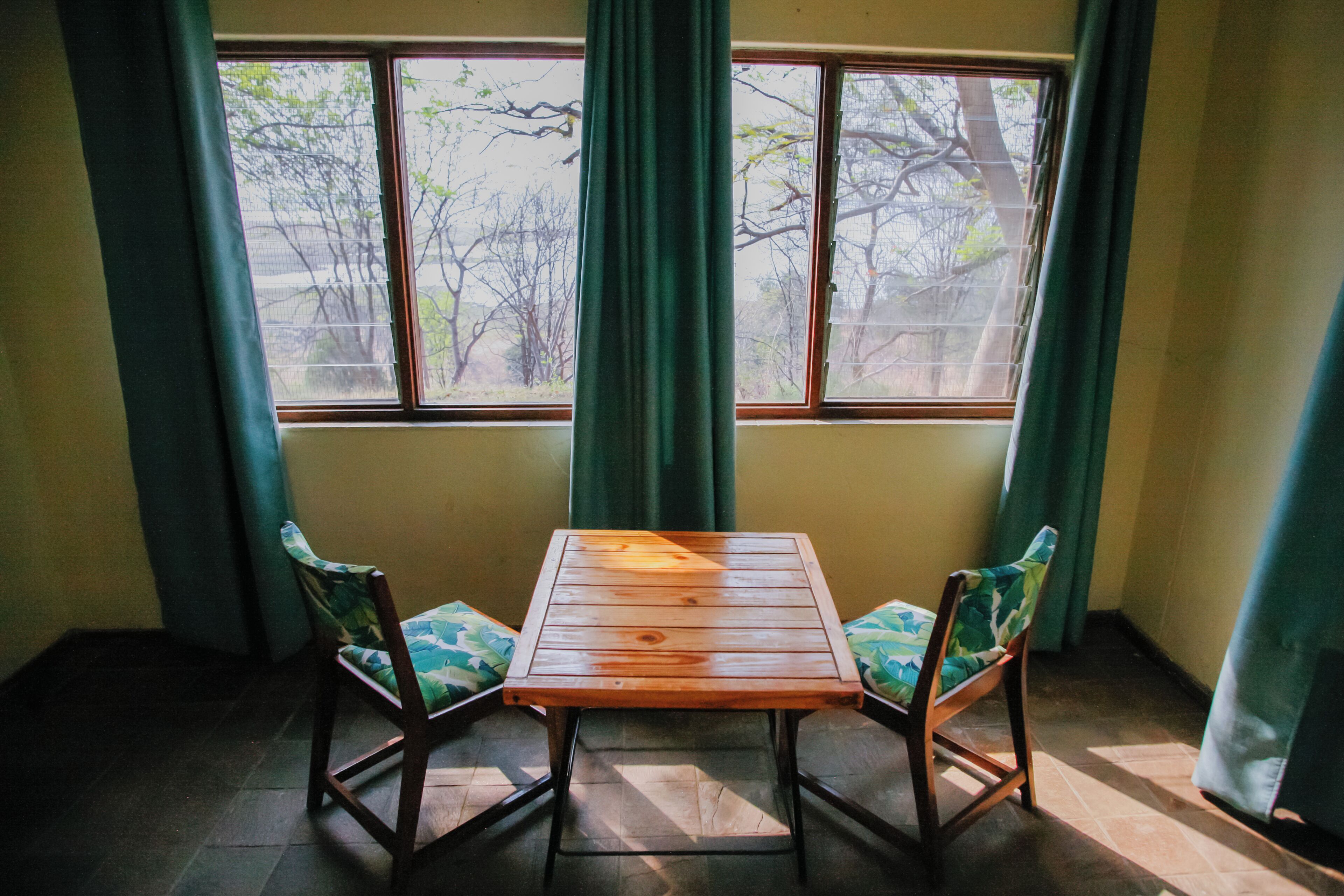 Habitación con una mesa de madera y dos sillas con cojines estampados, una ventana con cortinas y vista a árboles.