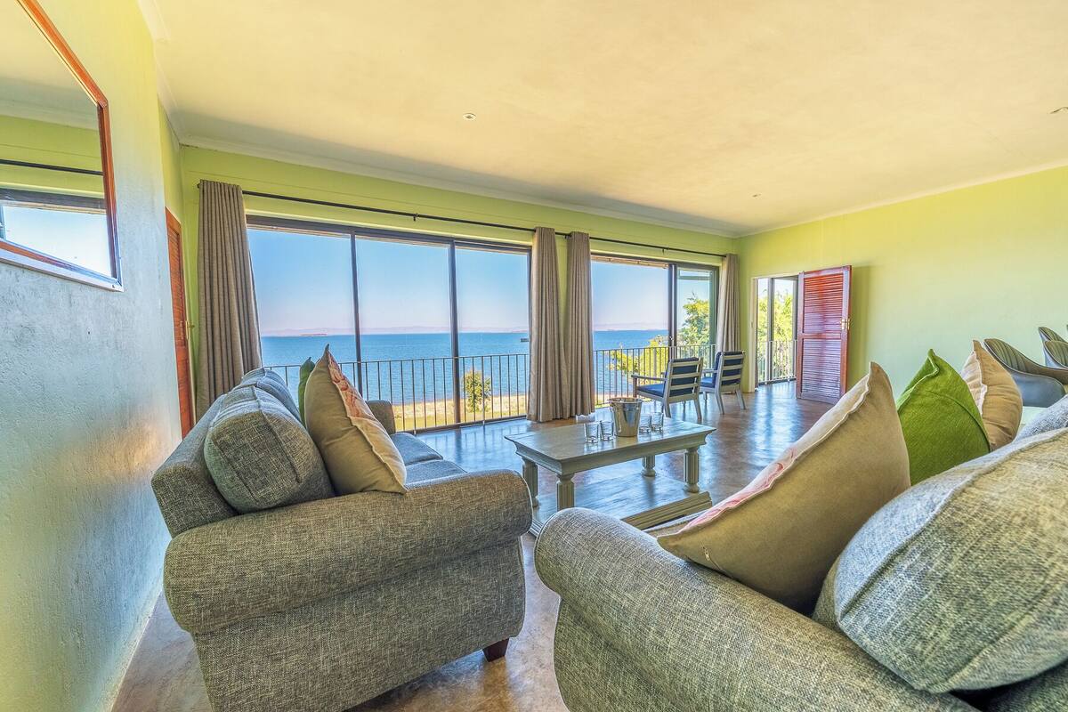 A spacious living area with a large window overlooking the sea, a dining table with chairs, and a balcony with a view of the ocean.