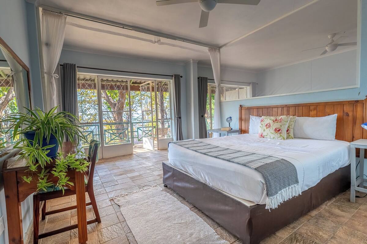 A spacious bedroom with a large bed, a wooden headboard, and a view of the outdoors through large windows.