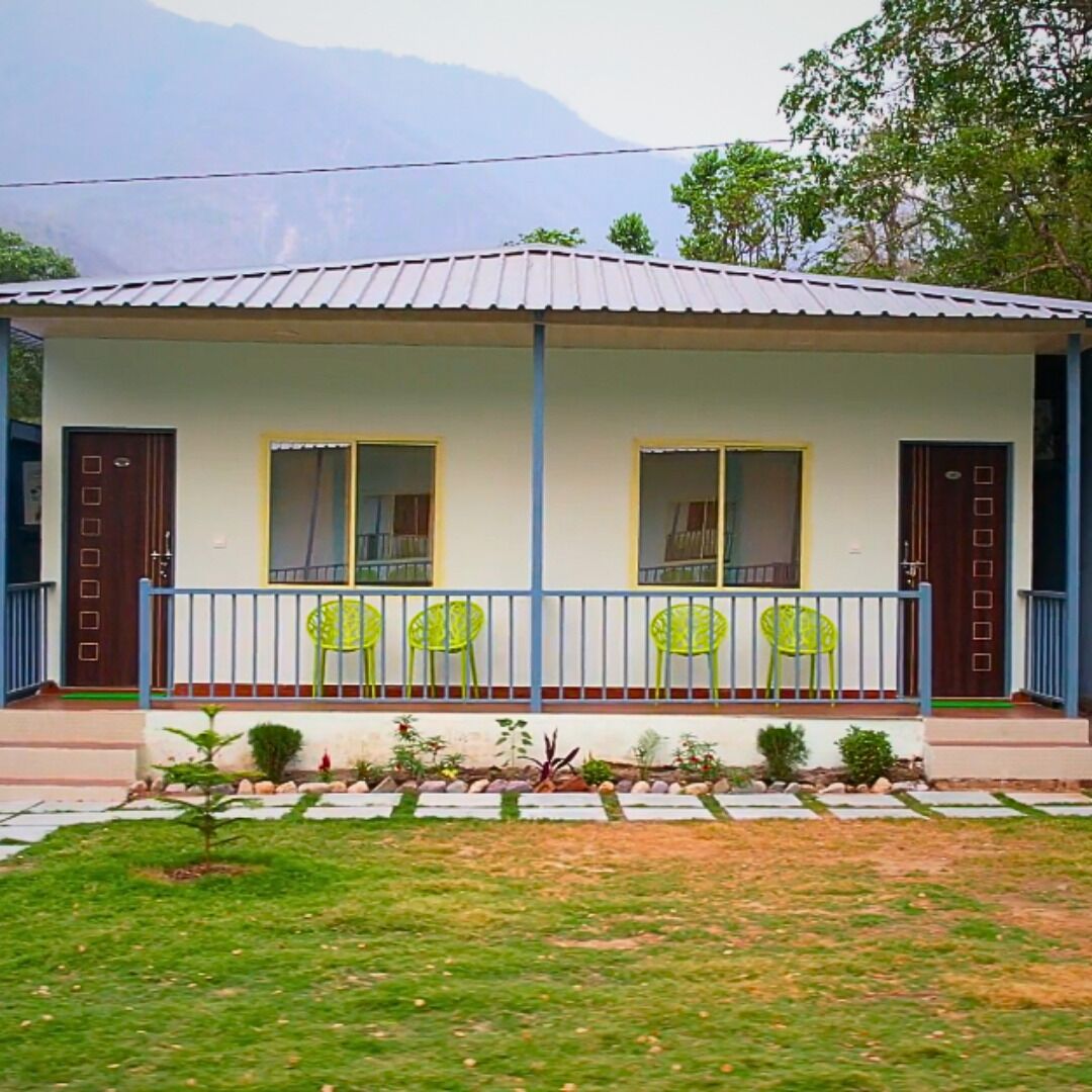 A single-story house with a flat roof, two windows, and a porch with green plastic chairs.