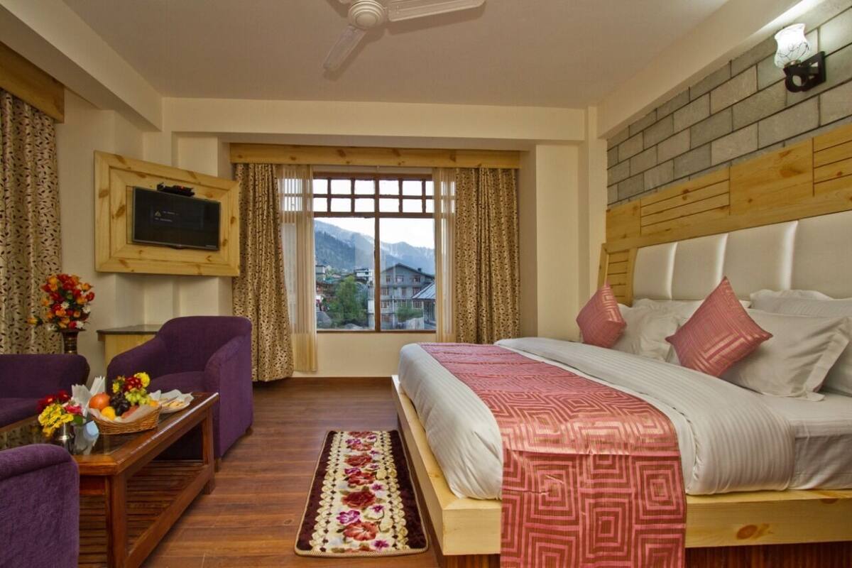A hotel room with a bed, a TV, a window with curtains, a wooden headboard, and a small table with flowers.