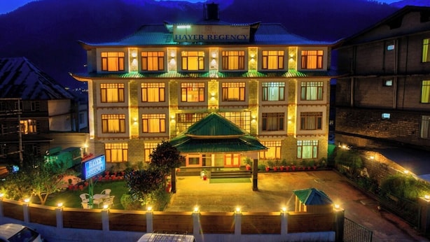 A multi-story hotel with a traditional architectural style, featuring a prominent entrance with a green roof and illuminated windows. - Hayer Regency (Manali)