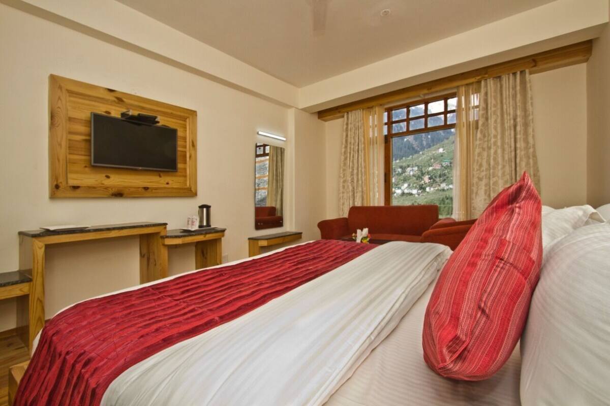 A hotel room with a large bed, a wooden desk, a television mounted on the wall, and a window with curtains.