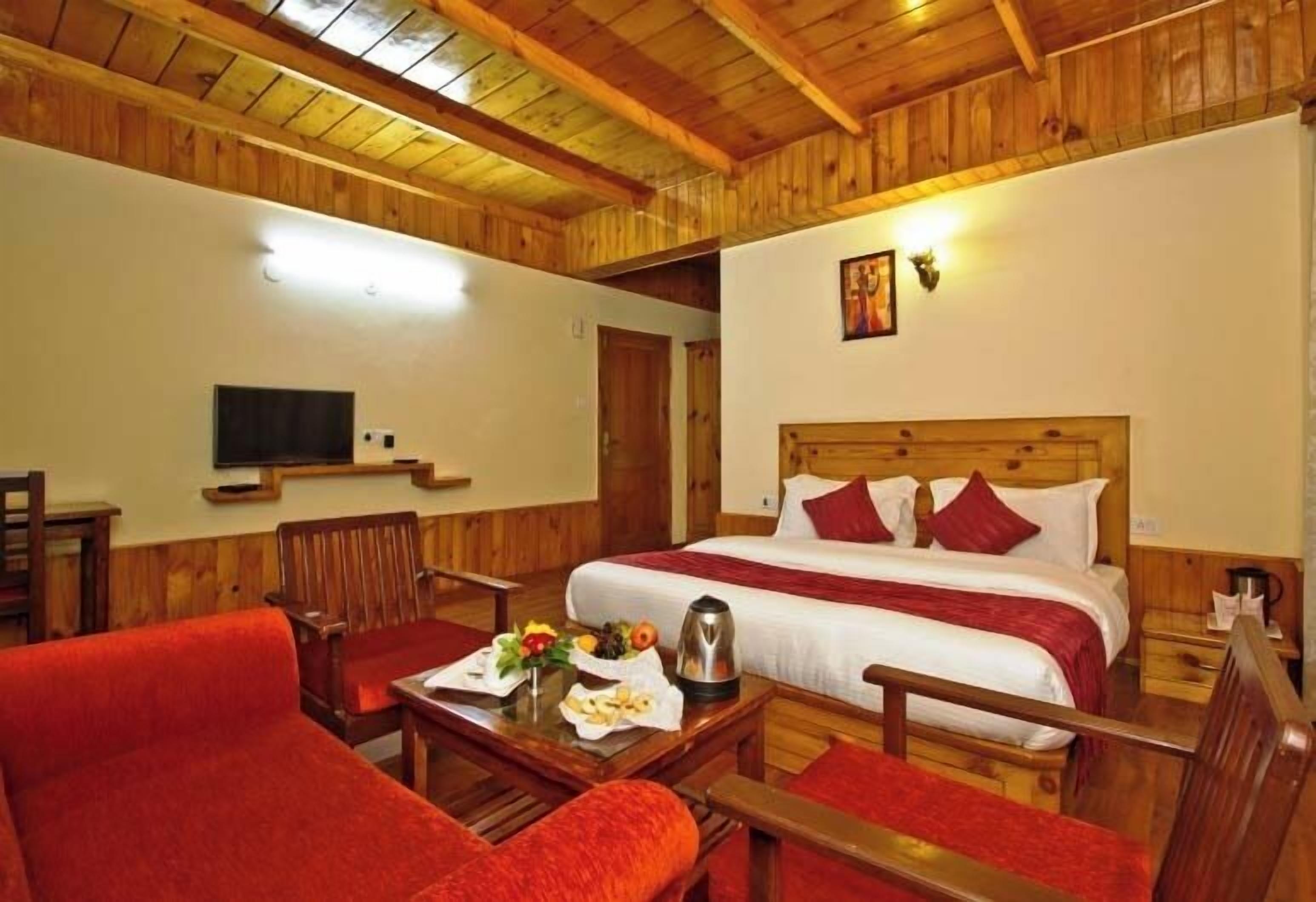 A wooden-themed hotel room with a bed, red upholstered chairs, a table with breakfast, and a television on the wall.