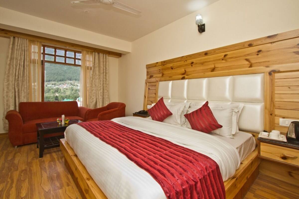 A bedroom with a wooden headboard, a large bed with red and white bedding, a red sofa, and a view of a mountainous landscape through the window.