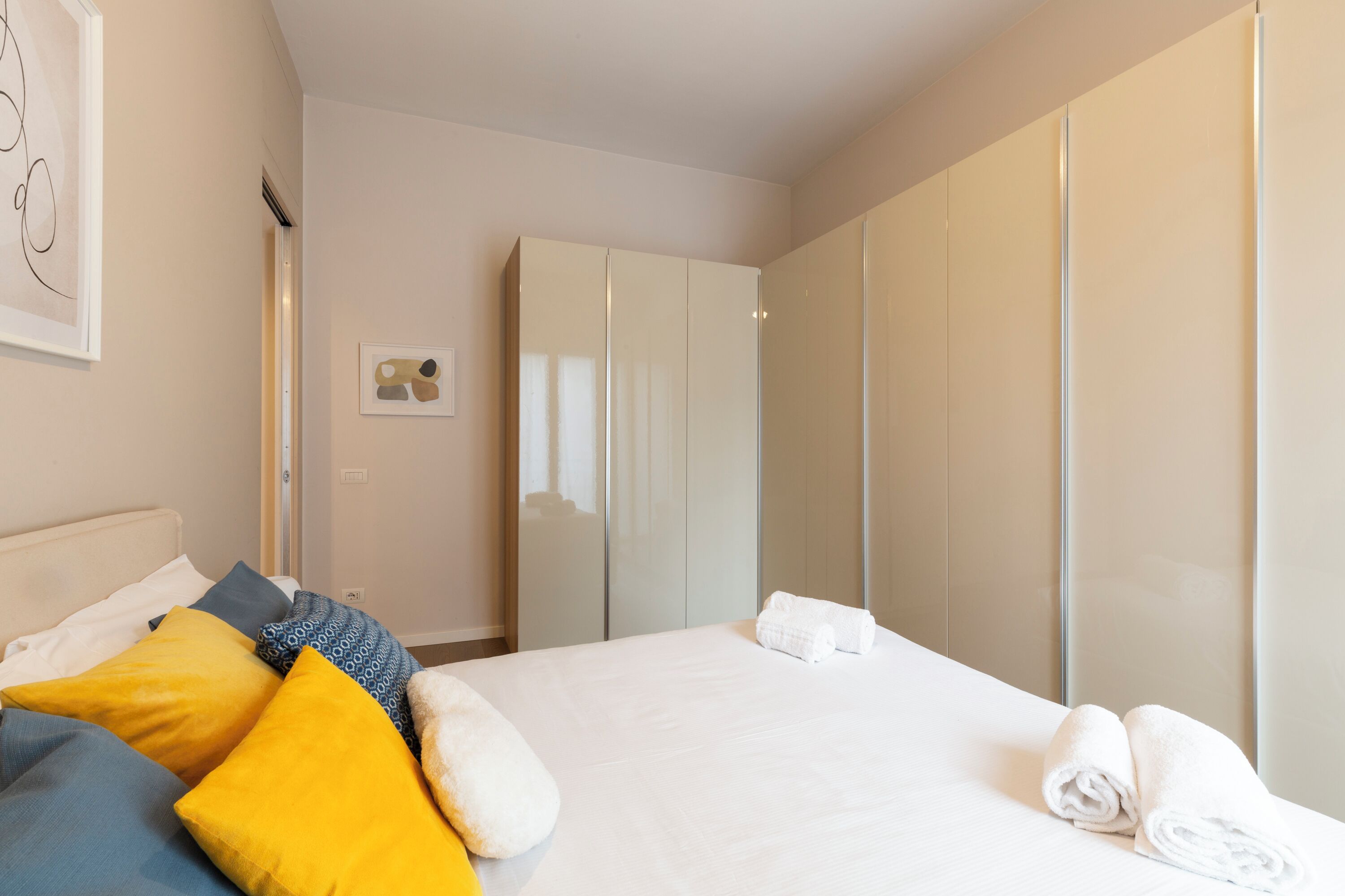 Een hotelkamer met een bed, kussens, een garderobe en een gekaderd schilderij aan de muur.