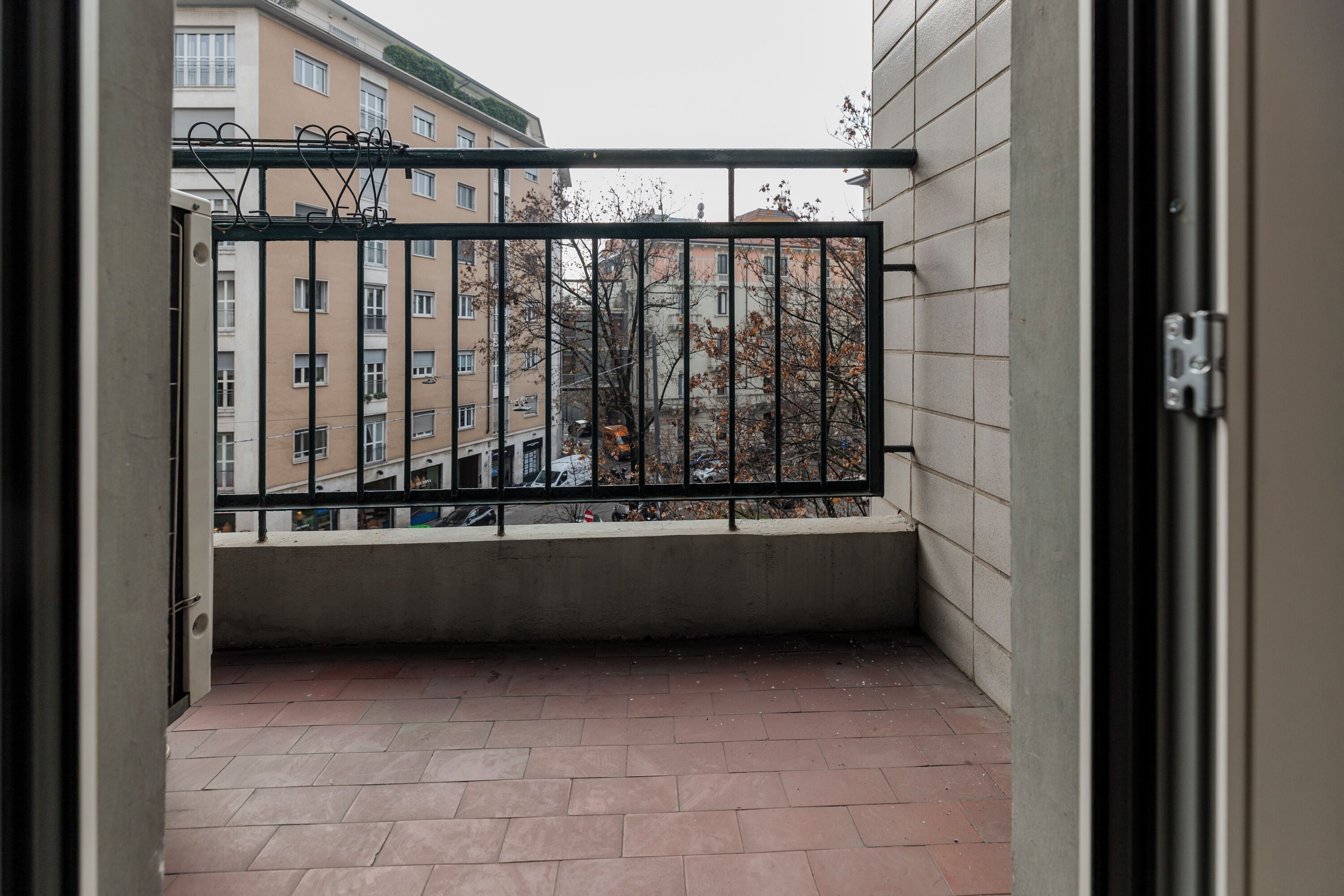 Een balkon met een metalen hekwerk, betegelde vloer en een uitzicht op gebouwen en bomen.