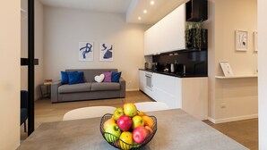 A modern living room with a grey sofa, a coffee table with a fruit bowl, and a kitchen area with a black countertop and white cabinets. - notaMi - Arena Modern Apartment (Milan)