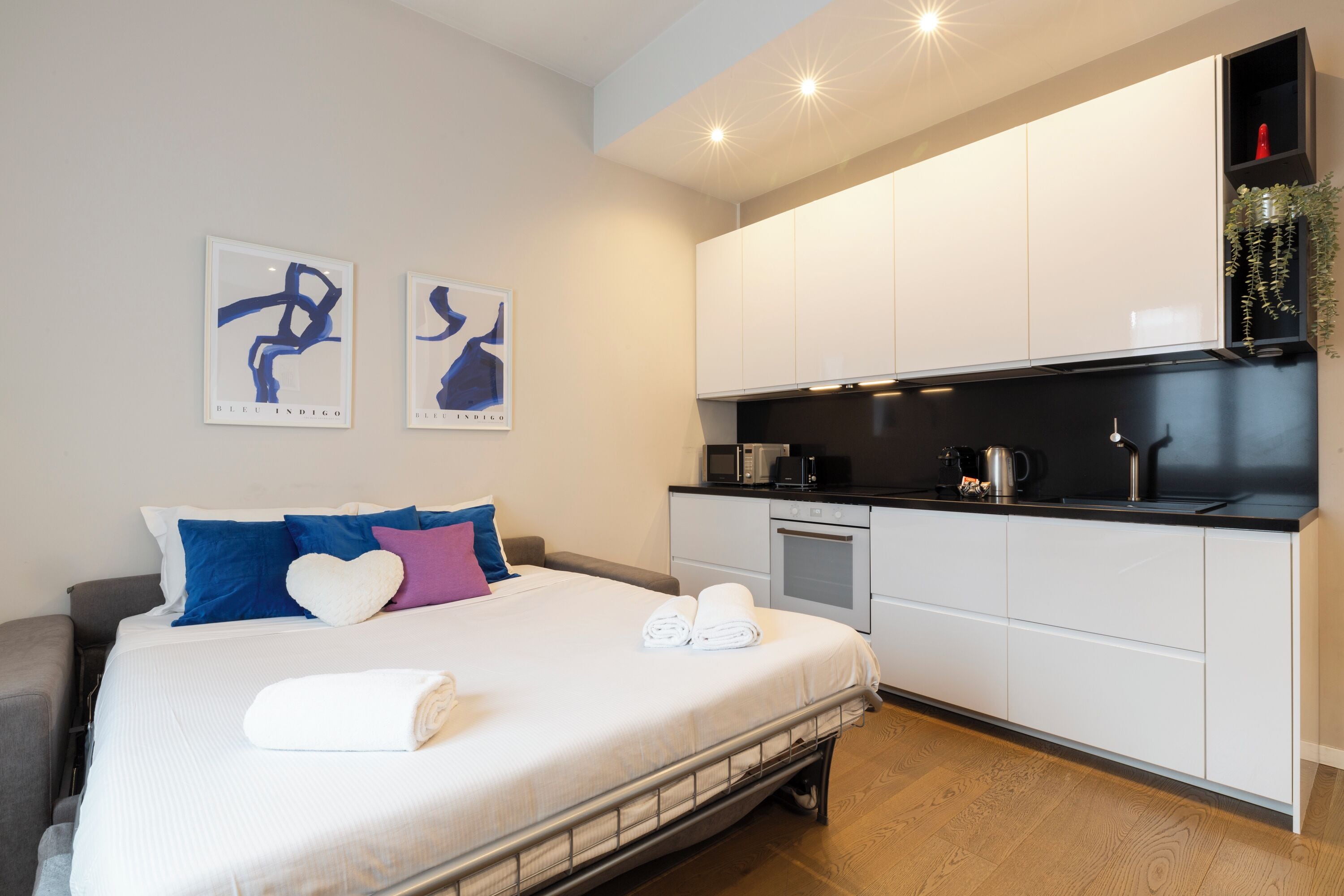 A modern hotel room with a bed, a kitchenette, and artwork on the wall.