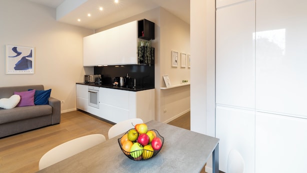 A modern kitchen with white cabinets, a black countertop, and a built-in oven. There is a dining table with a fruit bowl in the foreground. - notaMi - Arena Modern Apartment (Milan)