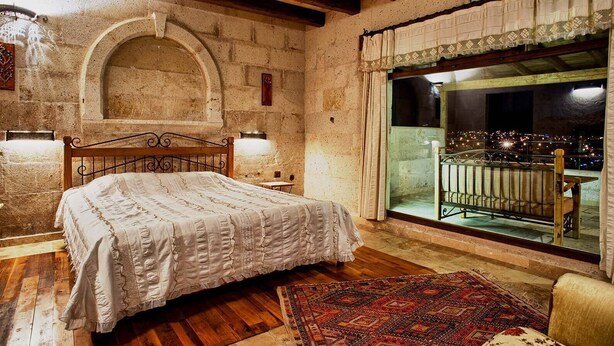 A bedroom with a wooden bed, a large window with a balcony, stone walls, and a rustic wooden ceiling. - Avanos Evi (Avanos)